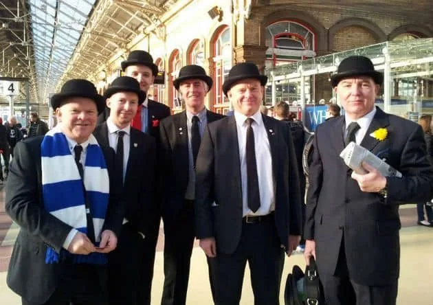 Group of supporters in bowlers and suits together at a station before the match.